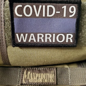 May include: A black patch with white text that reads "COVID-19 WARRIOR" and a blue stripe. The patch is attached to a green fabric bag.