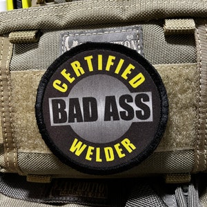Può includere: Una toppa ricamata nera e gialla con il testo "Certified Bad Ass Welder" su uno sfondo grigio.