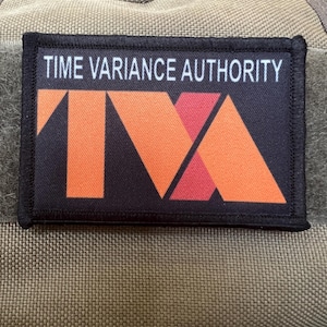 Puede incluir: Un parche negro con texto blanco que dice "TIME VARIANCE AUTHORITY" y un logotipo rojo y naranja con las letras "TVA" en el centro.