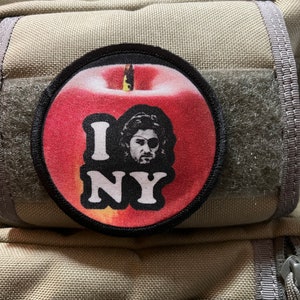 May include: A red apple with a black outline and a white "I NY" graphic. The apple has a black stem and a white leaf. A person's face is visible in the centre of the apple.