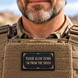 May include: A rectangular black morale patch with gold embroidered text that reads PLEASE SLOW DOWN I'M FROM THE 1900S, featuring a decorative gold border with ornate corner details, attached to a tan tactical vest with a camouflage shirt underneath.