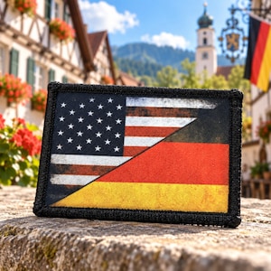 May include: A rectangular patch featuring a split design that combines the United States flag on the left with the German flag on the right. The patch has a thick black stitched border and a weathered, distressed texture across the surface.