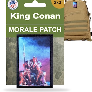 Könnte beinhalten: Ein 2x3 Zoll großer Morale-Patch mit dem Text "King Conan" und "Morale Patch". Der Patch zeigt einen Krieger mit einem Speer, der auf einem Thron sitzt. Der Patch ist an einem beigefarbenen Rucksack befestigt. Hergestellt in den USA.