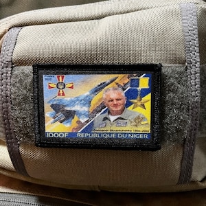 Puede incluir: Un parche negro con una imagen colorida de un avión de combate y un hombre con uniforme de piloto. El parche tiene el texto "1000F REPUBLIQUE DU NIGER" y "Oleksandr Oksanchenko 1980-2022".