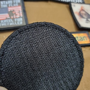 May include: A black, round, fabric patch with a textured surface. The patch is designed to be sewn onto clothing or other items.