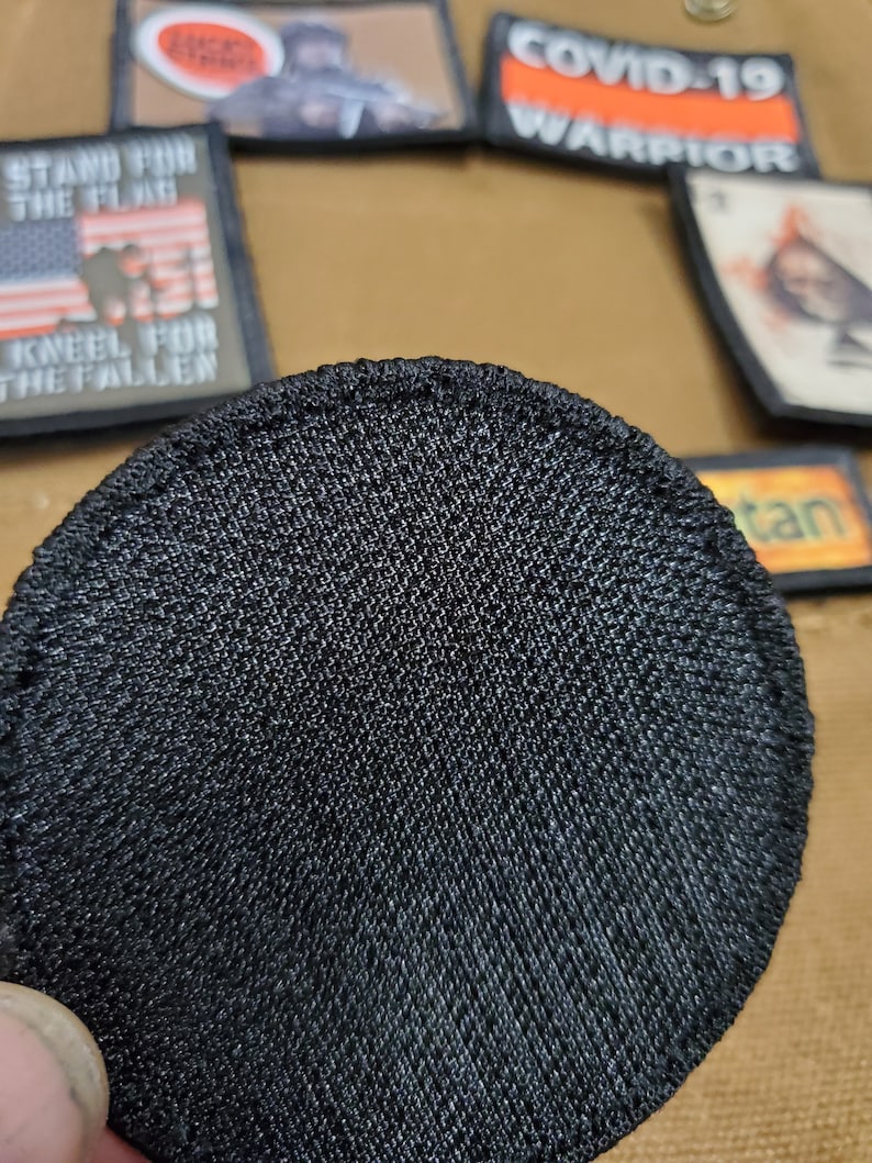May include: A black, round, fabric patch with a textured surface. The patch is designed to be sewn onto clothing or other items.