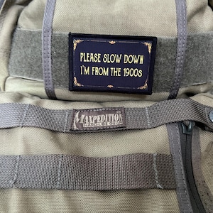 May include: A tan Maxpedition Hard-Use Gear backpack with a black patch that reads "Please Slow Down I'm From The 1900s".