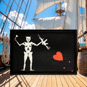 May include: A rectangular black patch featuring a white embroidered skeleton figure holding a spear in one hand and a rifle in the other. To the right of the skeleton, there is a red heart shape with three small red dots trailing below it.