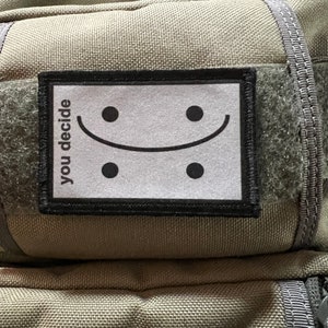May include: A green tactical backpack with a black patch that says "you decide" and a white smiley face with black eyes.