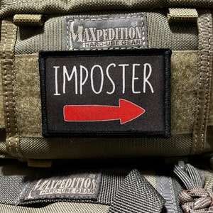 May include: A black patch with white text that reads "IMPOSTER" and a red arrow pointing to the right. The patch is attached to a green tactical bag with the Maxpedition logo.