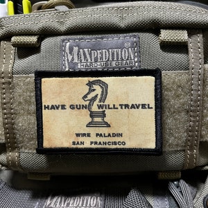 May include: A tan and black patch with the text "HAVE GUN WILL TRAVEL" and a chess piece graphic. The text "WIRE PALADIN SAN FRANCISCO" is below the graphic. The patch is attached to a green and tan bag with the text "MAXPEDITION HARD-USE GEAR" on it.