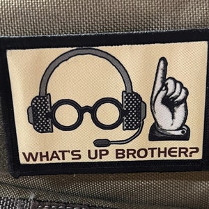 Puede incluir: Un parche bordado en blanco y negro con una imagen de dibujos animados de una persona con auriculares y gafas con el texto "WHAT'S UP BROTHER?"