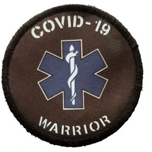 Op de afbeelding: Een ronde bruine patch met een wit ster-van-leven-symbool en de tekst "COVID-19 WARRIOR" in wit.