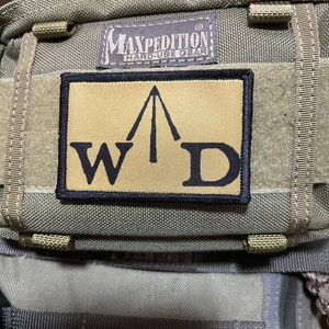 Op de afbeelding: Een beige en zwarte patch met de tekst "MAXPEDITION HARD-USE GEAR" en een zwarte driehoek met de letters "W" en "D" erin.