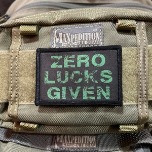 Puede incluir: Un parche verde y negro con el texto "ZERO LUCKS GIVEN" en una mochila Maxpedition Hard-Use Gear.