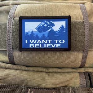 Pode incluir: Uma mochila bege com um remendo azul e branco que diz "I WANT TO BELIEVE" e uma imagem azul de um TARDIS voando sobre uma floresta. A mochila tem uma etiqueta preta que diz "MAXPEDITION HARD-USE GEAR".
