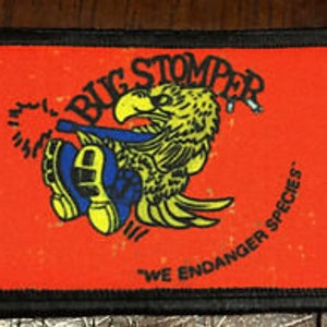 Pode incluir: Um remendo bordado vermelho com uma borda preta. O remendo apresenta um pássaro de desenho animado usando sapatos azuis e amarelos, pisando em um inseto. O texto "Bug Stomper" é escrito em um círculo ao redor do pássaro. O texto "We Endanger Species" é escrito abaixo do pássaro.