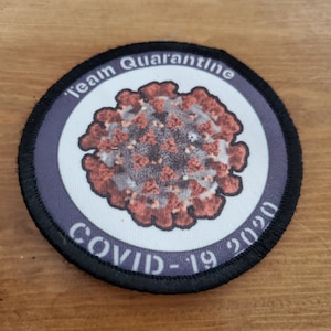 May include: A round, black, embroidered patch with a white background. The patch features a stylized image of the COVID-19 virus in red and brown. The text "Team Quarantine COVID-19 2020" is printed in black around the image.