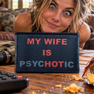 Puede incluir: Un parche rectangular de tela azul marino oscuro con el texto MY WIFE IS PSYCHOTIC en letras rojas y azul claro, gruesas y en bloque. El parche tiene un borde cosido y una superficie texturizada, presentando un mensaje claro y legible.