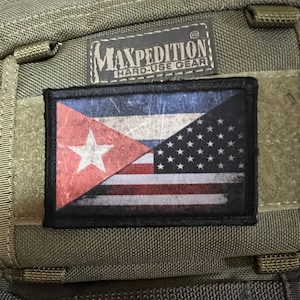 May include: A black patch with a distressed American flag and a Cuban flag superimposed on top of it. The patch is attached to a green tactical bag with the text "Maxpedition Hard-Use Gear" on the bag.