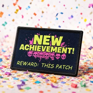May include: A rectangular black embroidered patch featuring the bright yellow text NEW ACHIEVEMENT! above a row of six pink skulls with a paint splatter effect. Below the skulls, the text REWARD: THIS PATCH is embroidered in yellow.