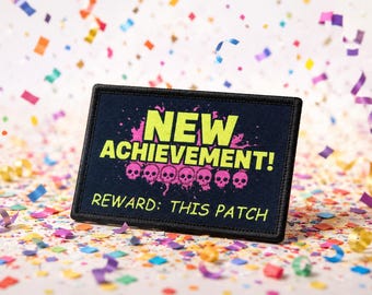 May include: A rectangular black embroidered patch featuring the bright yellow text NEW ACHIEVEMENT! above a row of six pink skulls with a paint splatter effect. Below the skulls, the text REWARD: THIS PATCH is embroidered in yellow.