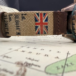 May include: A wristwatch with a weathered bronze-toned case and a green dial. The strap is made of tan canvas featuring a printed Union Jack flag and black geographic coordinates, accented with dark brown leather sections at the ends.