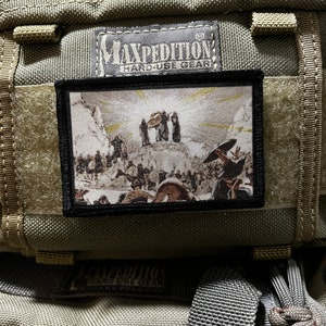 May include: A black and white patch with a scene of people walking towards a mountain with a bright light shining down. The patch is sewn onto a green tactical bag with the text "Maxpedition Hard-Use Gear" on the bag.