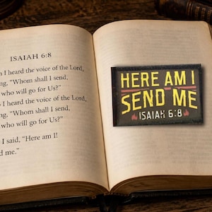 Op de afbeelding: Een rechthoekige zwarte patch met de gele tekst "HERE AM I SEND ME" en "ISAIAH 6:8" en kleine rode vlamiconen. De patch ligt op een opengeslagen bladzijde van een boek waarop de tekst van het bijbelvers Jesaja 6:8 te lezen is.