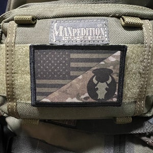 May include: A green tactical backpack with a black patch featuring a camouflage pattern and a bull's head logo. The patch also has the text "EXPEDITION HARD-USE GEAR".