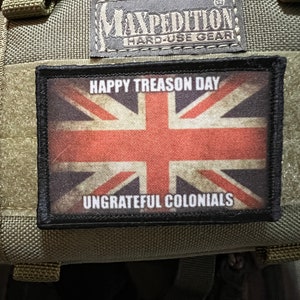 Puede incluir: Un parche negro con una bandera Union Jack roja, blanca y azul y el texto "Happy Treason Day" y "Ungrateful Colonials". El parche está unido a una bolsa Maxpedition verde.