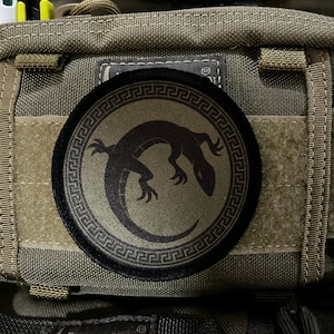 May include: A round patch with a black lizard design inside a Greek key border. The patch is olive green and black, and is attached to a tactical bag. The bag is a neutral color with multiple pockets and straps.