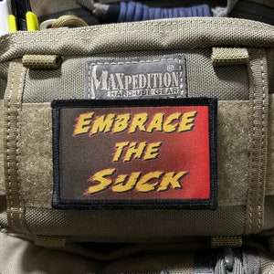 May include: A green Maxpedition Hard-Use Gear bag with a black and red patch that says "Embrace the Suck" in a comic book style font.