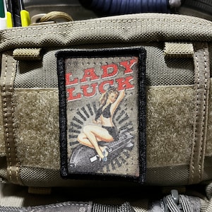 Puede incluir: Un parche marrón y negro con una ilustración de estilo vintage de una mujer en traje de baño sentada en un avión con el texto "Lady Luck" en letras rojas.