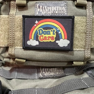 May include: A green tactical backpack with a black patch that says "Don't Care" with a rainbow and clouds. The backpack has the Maxpedition logo and the words "Hard-Use Gear" on it.