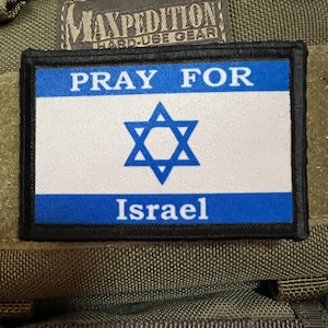 Puede incluir: Un parche negro con texto blanco que dice "Pray for Israel" y una estrella de David azul sobre fondo blanco. El parche está unido a un fondo de tela verde.