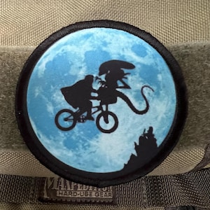 以下が含まれることがあります： 青い月の前で自転車に乗ったエイリアンのシルエットが描かれた白黒のパッチ。パッチは円形で、黒い縁取りがあります。