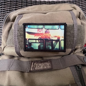 May include: A green military-style backpack with a black patch featuring a scene from the movie "Kill Bill". The patch shows a woman in a yellow jumpsuit standing on a bus with the words "Full Metal" written on the side. The backpack has a black strap and a black label that reads "Maxpedition Hard-Use Gear".