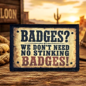 May include: A rectangular patch with a distressed beige background and a dark border. The text reads: BADGES? WE DON'T NEED NO STINKING BADGES! The lettering is in dark blue and maroon, featuring a small star icon between the lines of text.