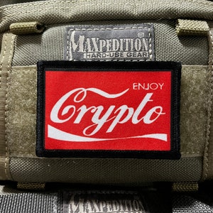 May include: A red and black patch with white text that says "Enjoy Crypto" on a green and gray fabric background. The patch is attached to a bag with the text "Maxpedition Hard-Use Gear" on it.