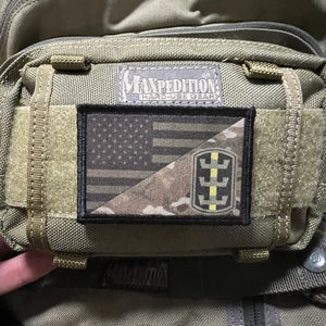 May include: A green tactical backpack with a black patch featuring a camouflage pattern and a yellow and black symbol. The patch is attached to the front of the backpack with velcro. The backpack has the text "Maxpedition Hard-Use Gear" printed on the front.