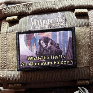 Könnte beinhalten: Ein schwarzer und gelber Patch mit einem Bild von Darth Vader und dem Text "What The Hell Is An Aluminum Falcon?" auf braunem Hintergrund.