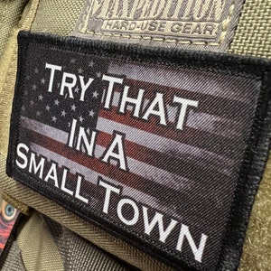 Pode incluir: Um remendo preto e branco com um design de bandeira americana e o texto "TRY THAT IN A SMALL TOWN". O remendo está preso a um fundo de tecido verde.