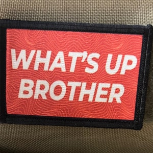 May include: A red and black patch with white text that reads "WHAT'S UP BROTHER" on a red background with a wavy pattern.