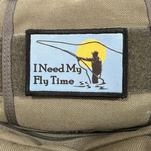 Könnte beinhalten: Eine braune Canvas-Tasche mit einem schwarzen Patch, auf dem "I Need My Fly Time" steht und eine Silhouette einer Person beim Fliegenfischen mit einer gelben Sonne im Hintergrund zu sehen ist.