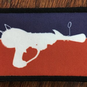 May include: A white silhouette of a toy ray gun on a red and blue fabric patch. The patch has a black border.