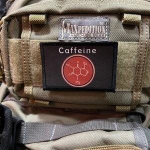 Puede incluir: Una mochila verde Maxpedition Hard-Use Gear con un parche negro que dice "Caffeine" y tiene una estructura química roja de cafeína dentro de un círculo.