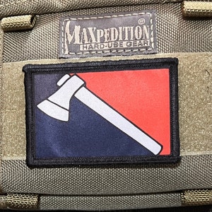 May include: A black and red patch with a white axe on a black background. The patch has a black border and the text "Maxpedition Hard-Use Gear" is visible above the patch.