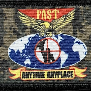 May include: A rectangular patch with a camouflage background. The patch features a red banner with the word "FAST" above a golden eagle. Below is a blue globe with a target and the words "ANYTIME ANYPLACE" on a yellow banner.