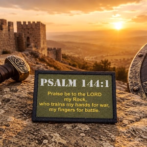 May include: A rectangular olive green patch featuring the white text PSALM 144:1 and the verse Praise be to the LORD my Rock, who trains my hands for war, my fingers for battle. The patch is positioned on a stone surface near a sword hilt and shield.